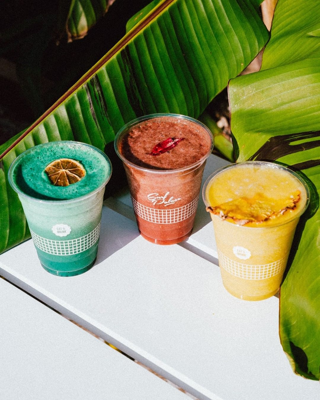 Colorful tropical smoothies in personalised smoothie cups with Surf Palace branding, featuring refreshing flavors and garnishes, set against lush green tropical leaves.