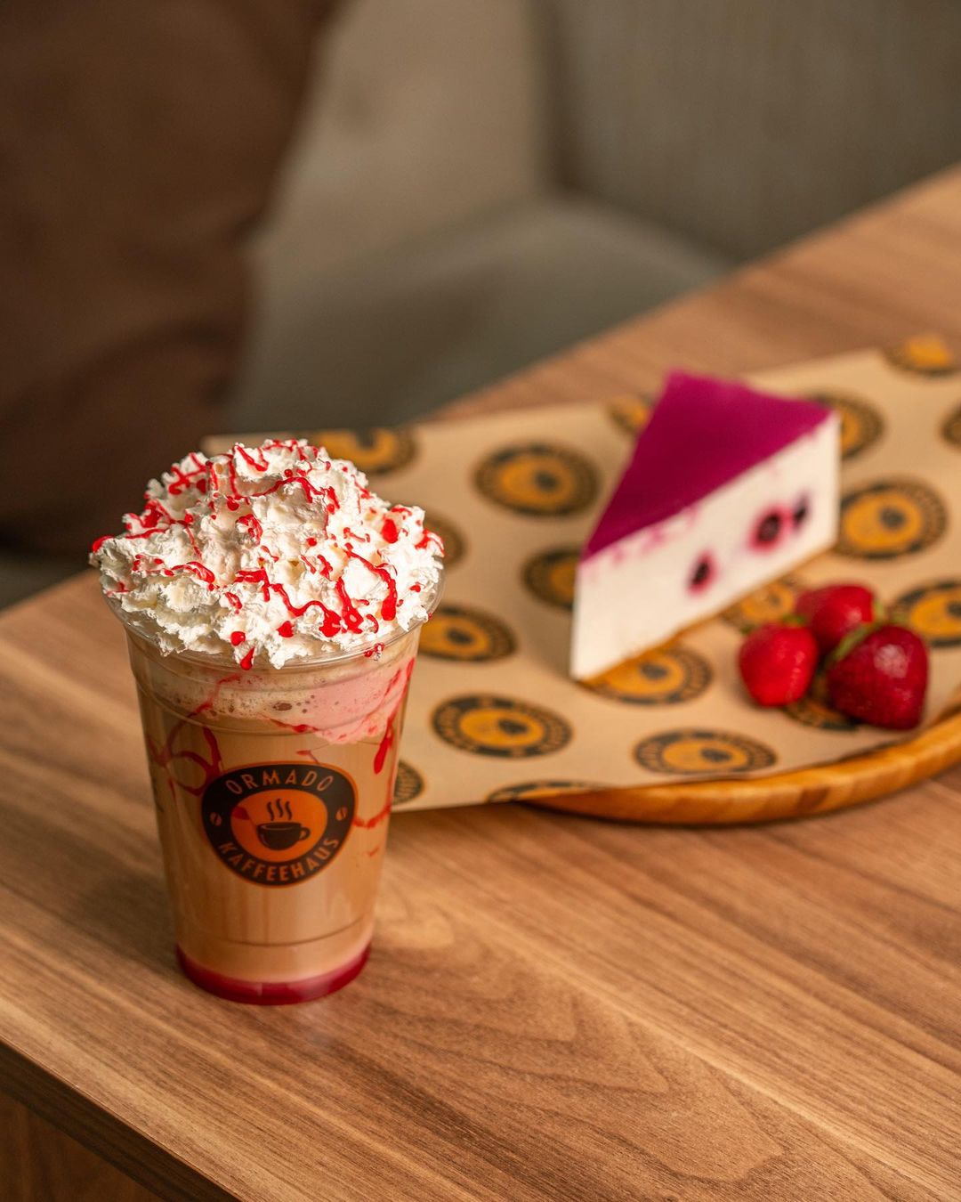 Iced coffee with whipped cream in a plastic cup with logo, served alongside berry cheesecake on a branded tray.