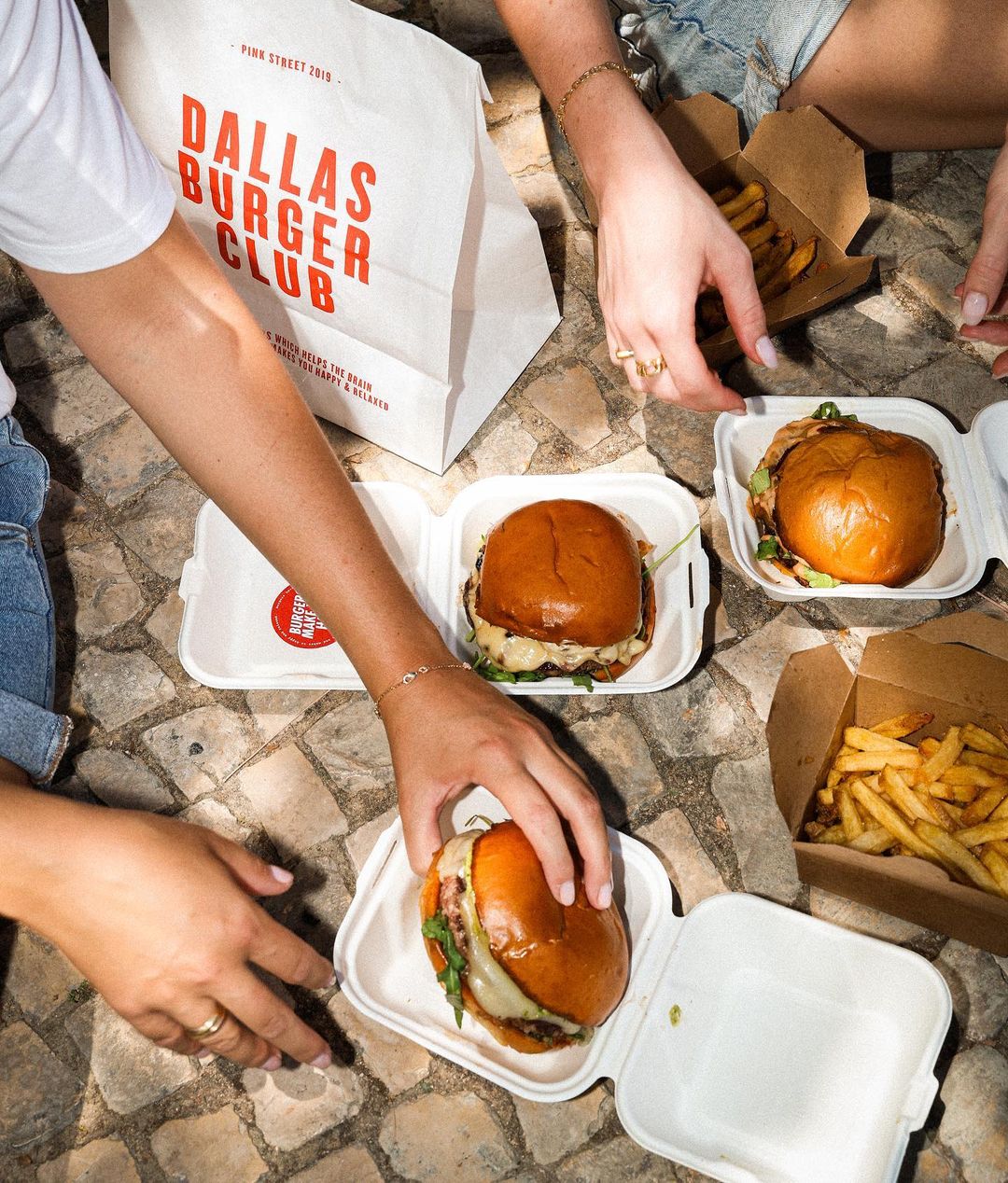 Group enjoying burgers and fries with a branded paper bag without handles from Dallas Burger Club in the background.