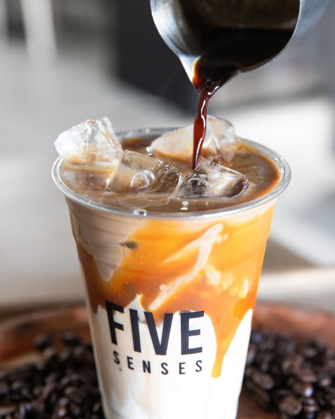 Pouring rich espresso into an iced latte served in a Personalised Milkshake Cup with Five Senses branding, creating a visually striking blend of coffee and milk.