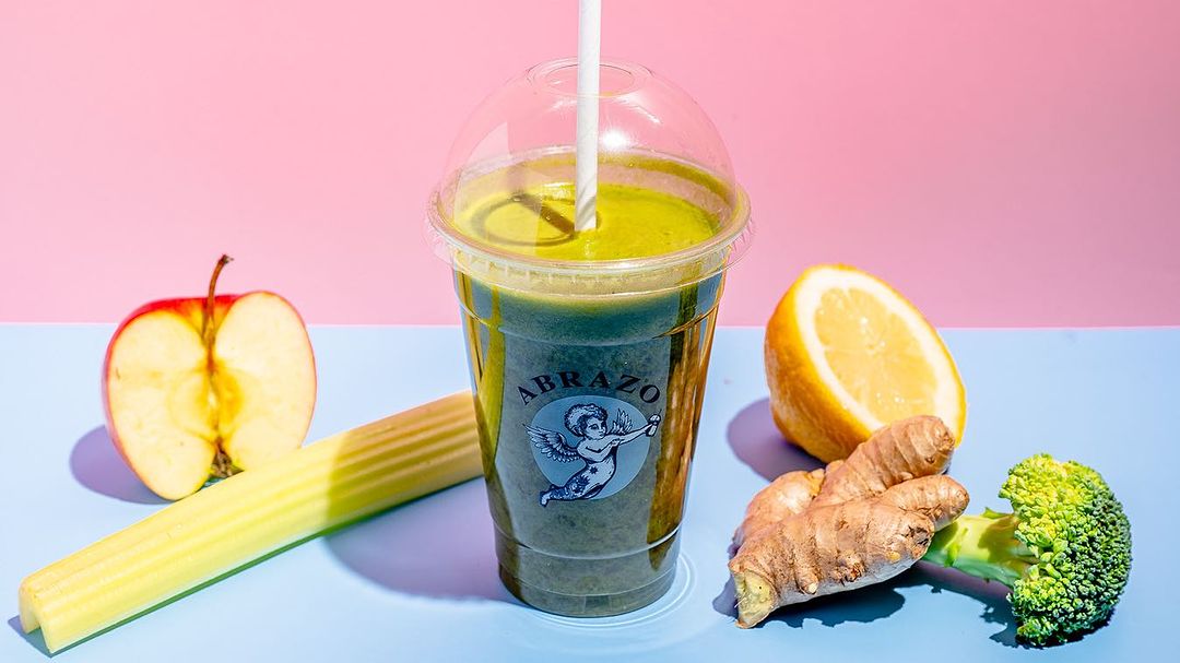 Healthy green smoothie in a personalised smoothie cup with Abrazo branding, accompanied by fresh ingredients like apple, celery, lemon, ginger, and broccoli for a vibrant, nutritious look.