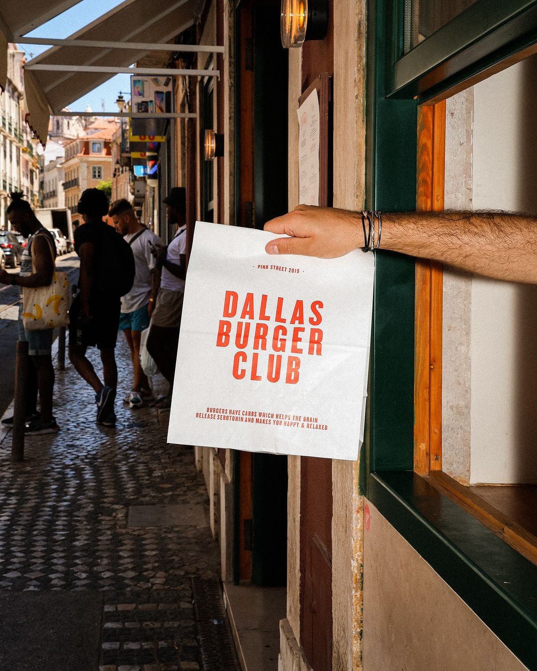 Branded takeaway bag from Dallas Burger Club, being handed out through a window onto a lively street. The bag prominently displays the Dallas Burger Club logo in bold red letters, making it a striking choice for burger takeaway packaging