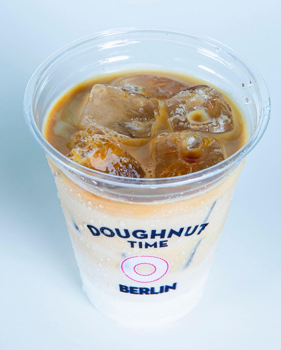 Iced coffee served in a branded plastic cup with logo, featuring the text 'Doughnut Time Berlin' printed on the cup, showcasing clear branding on a transparent plastic cup filled with ice.