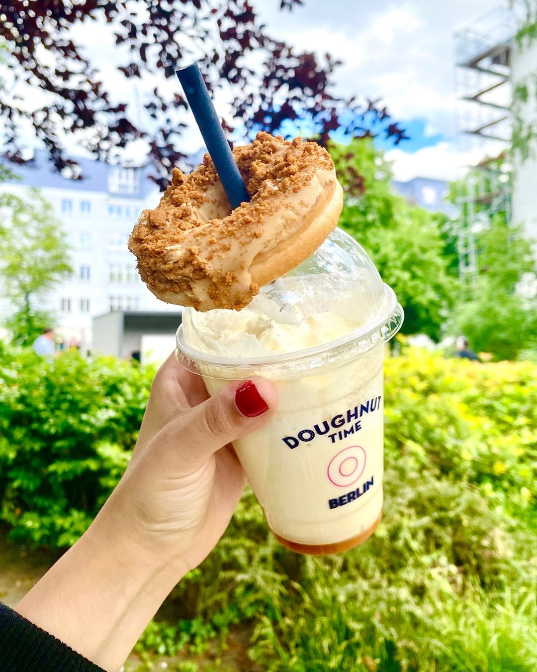 Hand holding a branded plastic cup with logo from 'Doughnut Time Berlin,' filled with a creamy beverage topped with whipped cream, and garnished with a donut on the straw.