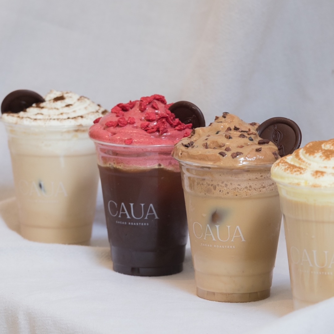 Four assorted iced drinks topped with whipped cream and garnishes in plastic cups with logo, showcasing a branded lineup for a café.