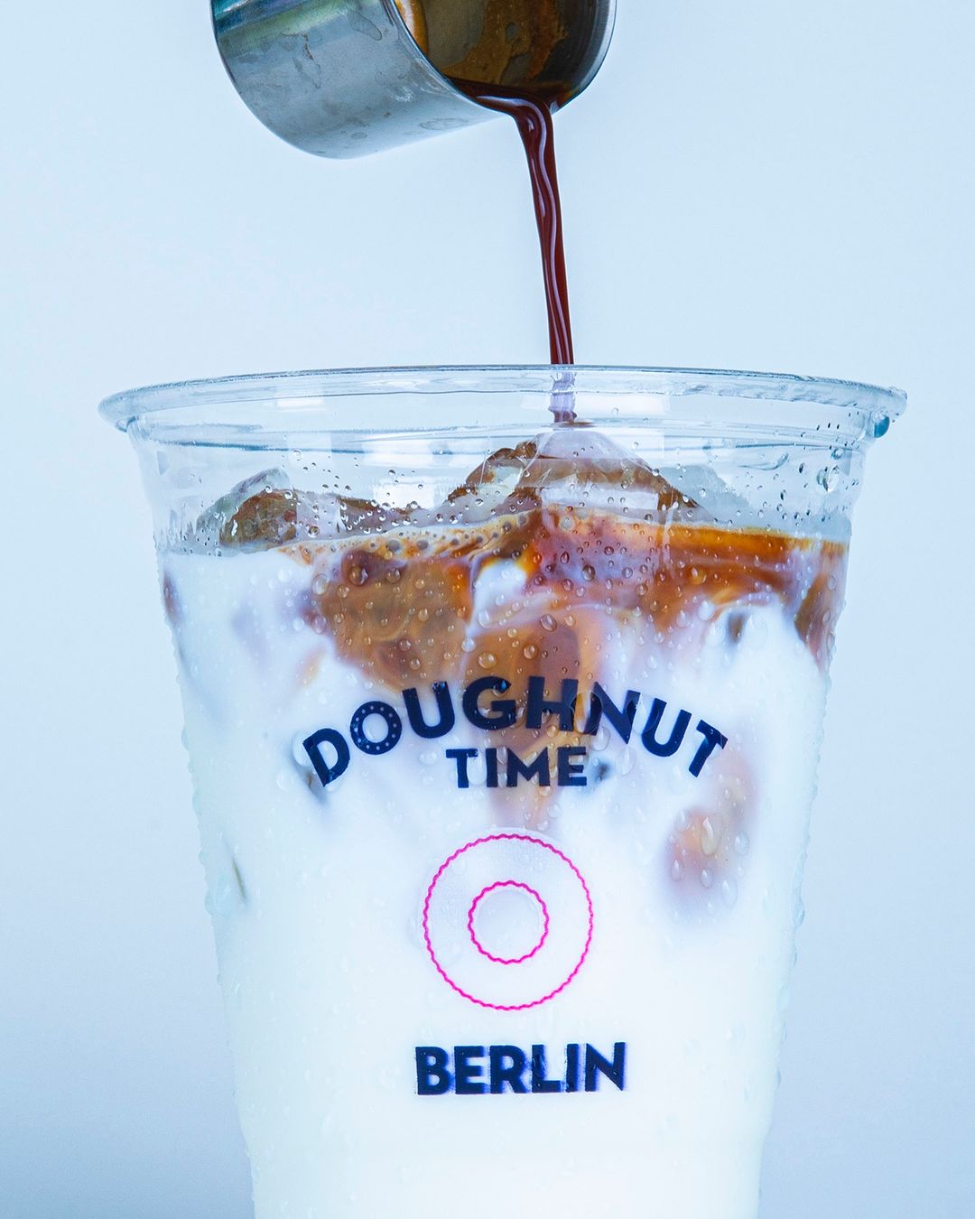 Branded plastic cup with logo from 'Doughnut Time Berlin,' filled with ice and milk, as espresso is poured in, creating a layered iced coffee effect in a clear plastic cup.