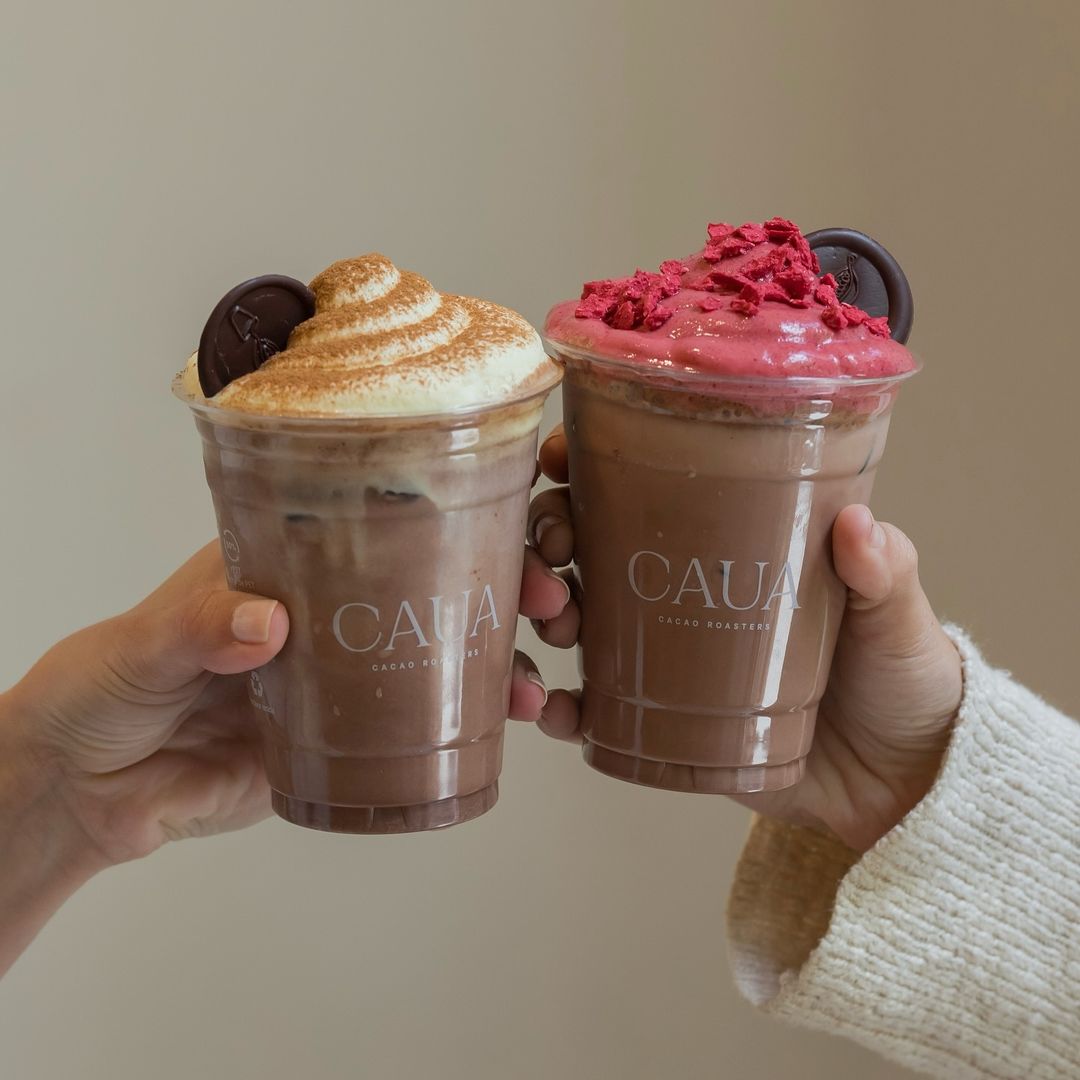 Two hands holding chocolate drinks topped with whipped cream and berries in plastic cups with logo, showcasing branded presentation for a café.