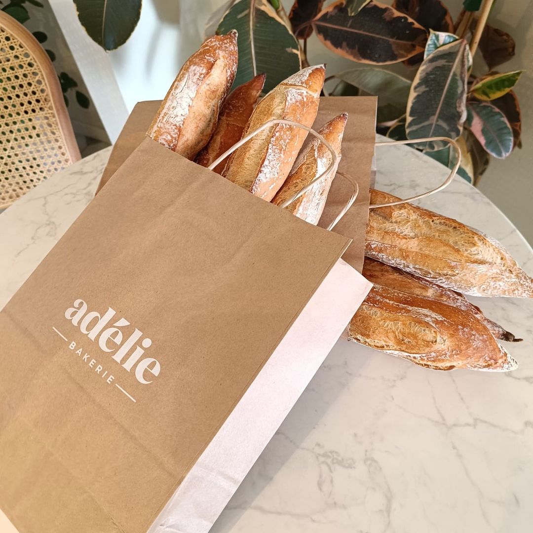 Branded takeaway bag from Adélie Bakery filled with freshly baked baguettes, placed on a marble table in a cozy setting. 