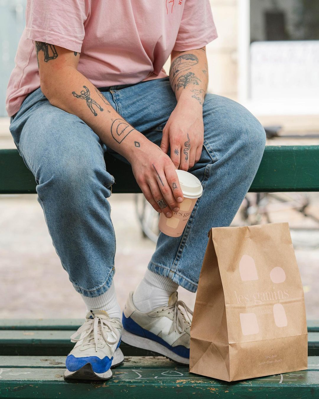 Person sitting on a bench holding a coffee cup with a block bottom paper bag from Les Gourmis placed beside them, featuring casual streetwear and tattoos.