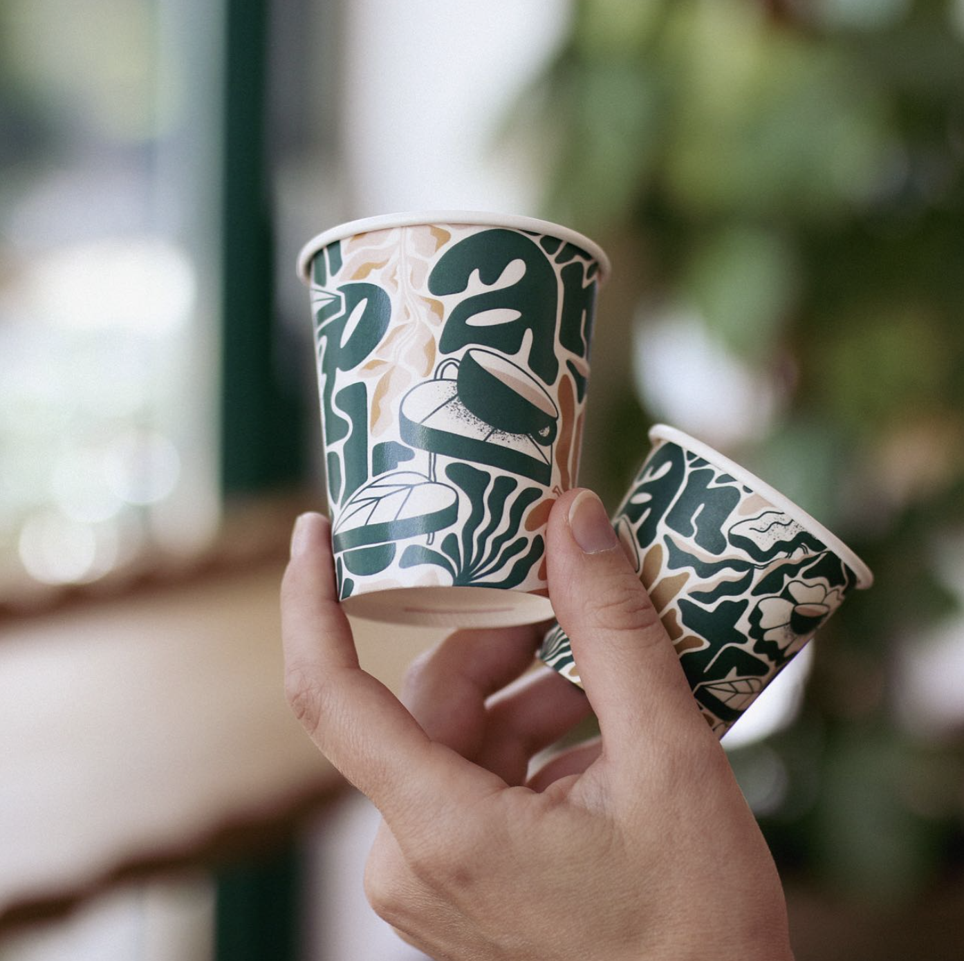 Two hands holding small custom paper cups with bold, abstract green and beige designs, ideal for coffee or cold beverages in a café setting.